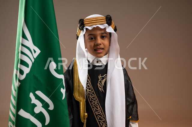 الوقوف بفخر بجانب العلم, يوم بدينا 1727 م، ارتداء أزياء المنطقة الوسطى في المناسبات الوطنية، ذكرى تأسيس الدولة السعودية 22 فبراير، بورتريه مقرب لولد عربي خليجي سعودي مبتسم يرتدي الغترة مع العقال الذهبي الشعبي والبشت ينظر إلى الكاميرا، خلفية بيج