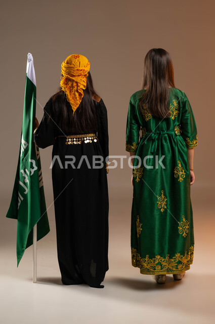 يوم بدينا 1727م, بورتريه من الخلف لفتاتين عربيتين خليجيتين سعوديتين ترتديان الزي الشعبي التقليدي في ذكرى تأسيس الدولة السعودية 22 فبراير، الوقوف باستقامة بجانب علم التاسيس بفخر, صورة الجسم بالطول الكامل, خلفية بيج