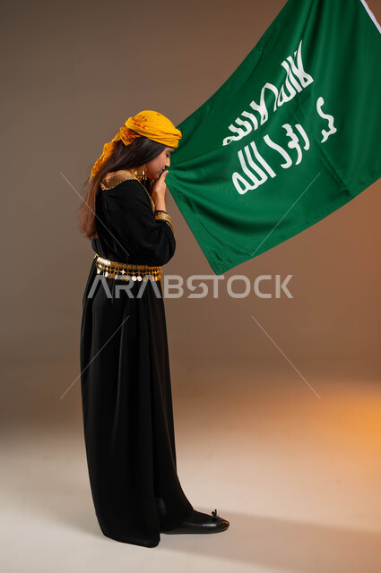 Anniversary of the founding of the first Saudi state on February 22, wearing the costumes of the central region on national holidays and occasions, portrait from the side of a Saudi Gulf Arab girl wearing a shawl and belt, proudly kissing the founding flag, Day of Our Life 1727 AD, full body portrait, beige background