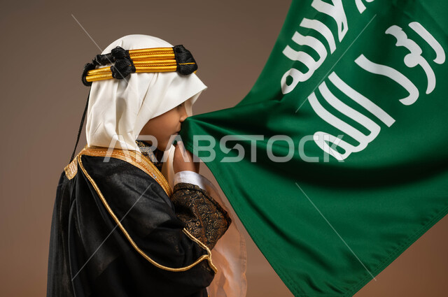 ارتداء أزياء المنطقة الوسطى في ذكرى تأسيس الدولة السعودية 22 فبراير، بورتريه مقرب من الجانب لولد عربي خليجي سعودي يرتدي البشت والعقال الذهبي يقوم بتقبيل علم التأسيس, إظهار مشاعر الفخر والاعتزاز بالوطن, يوم بدينا 1727م, تربيه الأبناء على حب الوطن، خلفية بيج