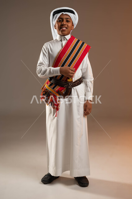 ذكرى تأسيس الدولة السعودية الأولى في 22 فبراير, ارتداء الزي التراثي الشعبي في المناسبات الوطنية, بورتريه لولد عربي خليجي سعودي مبتسم يرتدي الغترة والثوب التقليدي والشال الجنوبي يمسك بيده الخنجر, يوم بدينا 1727م,  صورة الجسم بالكامل, خلفية بيج