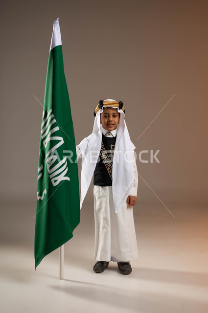 ارتداء زي المنطقة الشرقية في ذكرى تأسيس الدولة السعودية 22 فبراير، يوم بدينا 1727 م، بورتريه لولد عربي خليجي سعودي مبتسم يرتدي الثوب التقليدي والغترة مع العقال الذهبي يقف بجانب علم يوم التأسيس، الفخر والاعتزاز بالتاريخ السعودي، صورة الجسم بالكامل، خلفية بيج