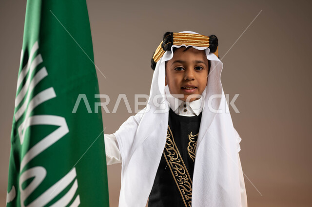 ارتداء الأزياء الشعبية في المناسبات الوطنية، ذكرى تأسيس الدولة السعودية 22 فبراير، يوم بدينا 1727 م، بورتريه مقرب لولد عربي خليجي سعودي مبتسم يرتدي الثوب التقليدي والغترة مع العقال الذهبي يقف بجانب علم يوم التأسيس، الفخر والاعتزاز بالتاريخ السعودي، خلفية بيج