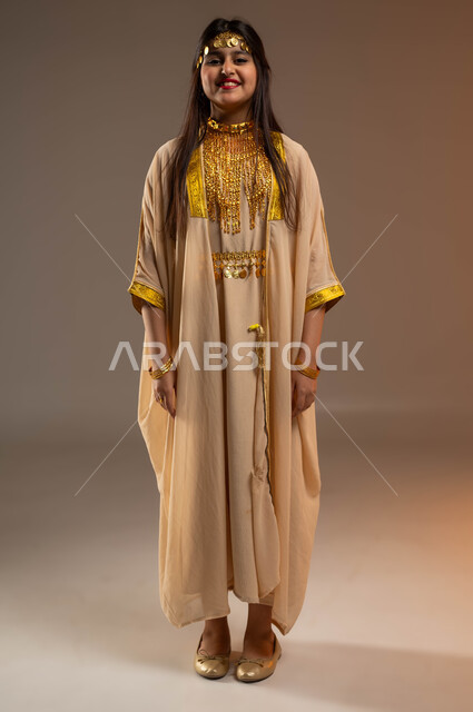 يوم بدينا 1727 م، ذكرى تأسيس الدولة السعودية 22 فبراير، ارتداء أزياء المنطقة الشرقية في المناسبات الوطنية، بورتريه لفتاة عربية خليجية سعودية مبتسمة ترتدي النشل تنظر إلى الكاميرا بإيماءات السرور والسعادة، التزين بالحلي والمجوهرات، صورة الجسم بالكامل، خلفية بيج
