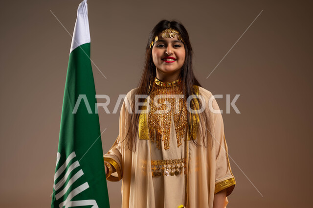 الفخر والاعتزاز بإنجازات البلاد، ذكرى تأسيس الدولة السعودية 22 فبراير، يوم بدينا 1727 م، التزيين بالحلي والمجوهرات، ارتداء أزياء المنطقة الشرقية في المناسبات الوطنية، بورتريه مقرب لفتاة عربية خليجية سعودية مبتسمة ترتدي النشل تقف بجانب علم يوم التأسيس، خلفية بيج