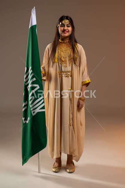 ذكرى تأسيس الدولة السعودية 22 فبراير، ارتداء أزياء المنطقة الشرقية في المناسبات الوطنية، بورتريه لفتاة عربية خليجية سعودية مبتسمة ترتدي النشل تقف بجانب علم يوم التأسيس، التزين بالحلي والمجوهرات الذهبية، يوم بدينا 1727 م، صورة الجسم بالكامل، خلفية بيج