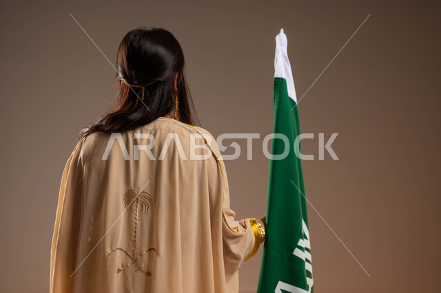 الاعتزاز والفخر بإنجازات البلاد، ذكرى تأسيس الدولة السعودية 22 فبراير، يوم بدينا 1727 م، التزيين بالحلي والمجوهرات، ارتداء أزياء المنطقة الشرقية في المناسبات الوطنية، بورتريه من الخلف لفتاة عربية خليجية سعودية ترتدي النشل تقف بجانب علم يوم التأسيس، خلفية بيج