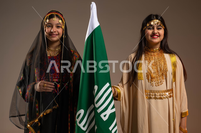 ارتداء أزياء المنطقة الشرقية والوسطى في المناسبات والأعياد الوطنية، يوم بدينا 1727 م، ذكرى تأسيس الدولة السعودية الأولى 22 فبراير، الوقوف باستقامة بجانب علم يوم التأسيس، بورتريه مقرب لفتاتين عربيتين خليجيتين سعوديتين مبتسمتين ترتديان زي شعبي تقليدي، خلفية بيج