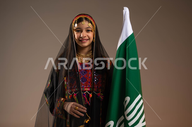 ذكرى تأسيس الدولة السعودية الأولى 22 فبراير، يوم بدينا 1727 م، بورتريه مقرب لفتاة عربية خليجية سعودية مبتسمة ترتدي الهامة والبشت والكرتة تقف بجانب علم يوم التأسيس تنظر الى الكاميرا، ارتداء أزياء المنطقة الوسطى، الفخر بالتراث التاريخي السعودي، خلفية بيج