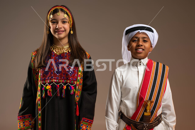 النظر الى الكاميرا بتعابير السرور، ذكرى تأسيس الدولة السعودية الأولى 22 فبراير، يوم بدينا 1727 م، بورتريه مقرب لولد عربي خليجي سعودي يرتدي الثوب والغترة مع الشال الجنوبي يقف بجانب فتاة مبتسمة ترتدي الهامة والكرتة، الفخر والاعتزاز بإنجازات الوطن، خلفية بيج