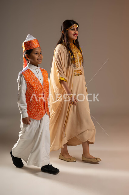 إيماءات التباهي والفخر بإنجازات الوطن، يوم بدينا 1727 م، ذكرى تأسيس الدولة السعودية الأولى 22 فبراير، ارتداء الأزياء الشعبية في المناسبات الوطنية، بورتريه لولد عربي خليجي سعودي يرتدي السديري يمشي بجانب فتاة ترتدي النشل التقليدي، صورة الجسم بالكامل، خلفية بيج