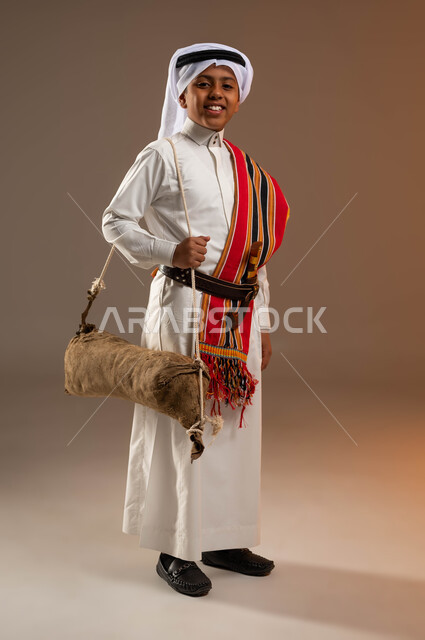 استخدام الادوات الشعبية، ارتداء أزياء المنطقة الجنوبية في يوم بدينا 1727 م، بورتريه لولد عربي خليجي سعودي يرتدي الثوب والغترة والشال الجنوبي يحمل قربة الماء للسقي ينظر إلى الكاميرا، ذكرى تأسيس الدولة السعودية الأولى 22 فبراير، صورة الجسم بالكامل، خلفية بيج