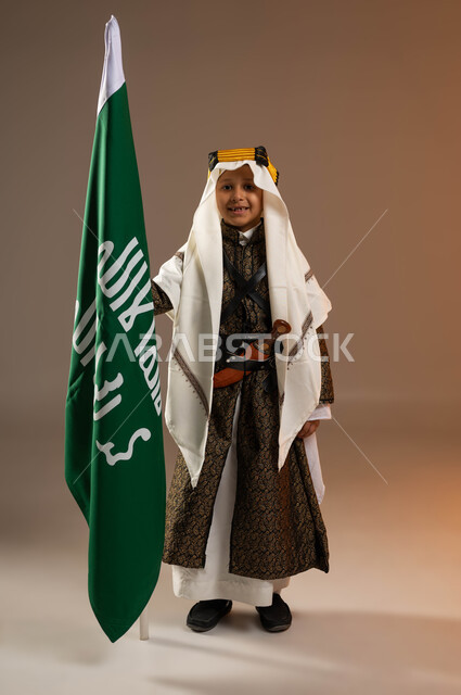 يوم بدينا 1727 م، النظر إلى الكاميرا بإيماءات الفخر، ارتداء أزياء المنطقة الوسطى في المناسبات الوطنية، بورتريه لولد عربي خليجي سعودي مبتسم يرتدي الدقلة والخنجر والعقال التقليدي الشعبي يقف بجانب علم تأسيس الدولة السعودية 22 فبراير، صورة لكامل الجسم، خلفية بيج
