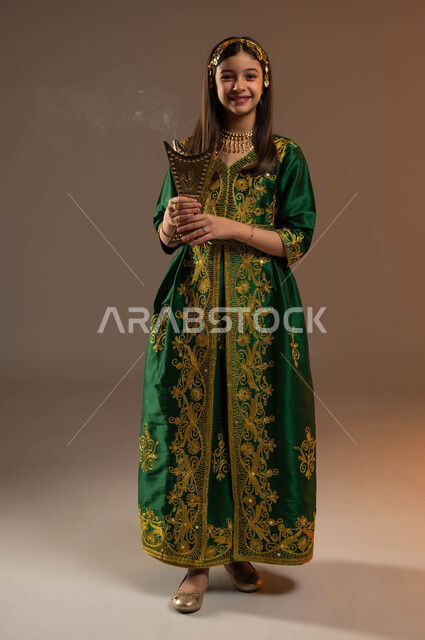 التطيب بالعود والبخور، يوم بدينا 1727 م، ذكرى تأسيس الدولة السعودية 22 فبراير، ارتداء أزياء المنطقة الوسطى، بورتريه لفتاة عربية خليجية سعودية مبتسمة ترتدي المقطع والحزام تحمل بيدها المبخرة تنظر إلى الكاميرا بإيماءات السرور والسعادة، صورة لكامل الجسم، خلفية بيج