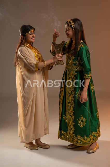 ارتداء أزياء المنطقة الشرقية والوسطى في الأعياد والمناسبات الوطنية، يوم بدينا 1727 م، ذكرى تأسيس الدولة السعودية الأولى 22 فبراير، بورتريه لفتاتين عربيتين خليجيتين سعوديتين مبتسمتين تقومان بالتعطر والتطيب بالعود والبخور السعودي، صورة لكامل الجسم، خلفية بيج