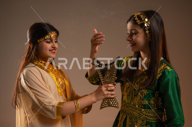 يوم بدينا 1727 م، ارتداء أزياء المنطقة الشرقية والوسطى في الأعياد والمناسبات الوطنية، ذكرى تأسيس الدولة السعودية الأولى 22 فبراير، بورتريه مقرب لفتاتين عربيتين خليجيتين سعوديتين مبتسمتين تقومان بالتعطر والتطيب بالعود والبخور السعودي، خلفية بيج