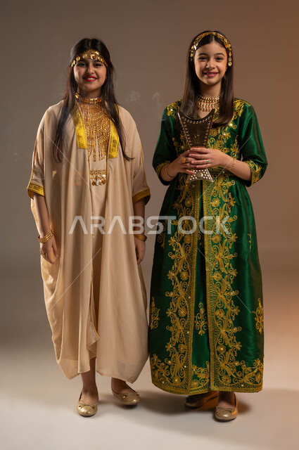 التطيب بالعود والبخور، ارتداء أزياء المنطقة الشرقية والوسطى في الأعياد الوطنية، يوم بدينا 1727 م، ذكرى تأسيس الدولة السعودية الأولى 22 فبراير، بورتريه لفتاتين عربيتين خليجيتين سعوديتين مبتسمتين تنظران إلى الكاميرا بتعابير السرور، صورة لكامل الجسم، خلفية بيج