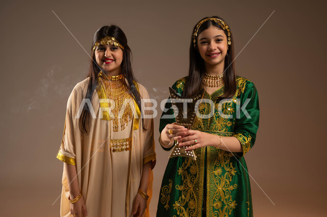 التطيب والتعطر بالعود والبخور، ارتداء أزياء المنطقة الشرقية والوسطى في المناسبات والأعياد الوطنية، يوم بدينا 1727 م، ذكرى تأسيس الدولة السعودية الأولى 22 فبراير، بورتريه لفتاتين عربيتين خليجيتين سعوديتين مبتسمتين تنظر إلى الكاميرا بتعابير السرور، خلفية بيج