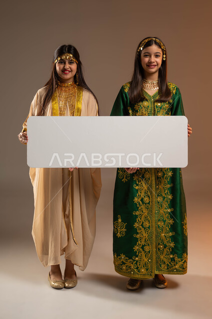 ذكرى تأسيس الدولة السعودية الأولى 22 فبراير، النظر إلى الكاميرا بإيماءات السرور، يوم بدينا 1727 م، ارتداء أزياء المنطقة الشرقية والوسطى، بورتريه لفتاتين عربيتين خليجيتين سعوديتين مبتسمتين تحملان موك أب أبيض فارغ كبير، صورة لكامل الجسم، خلفية بيج