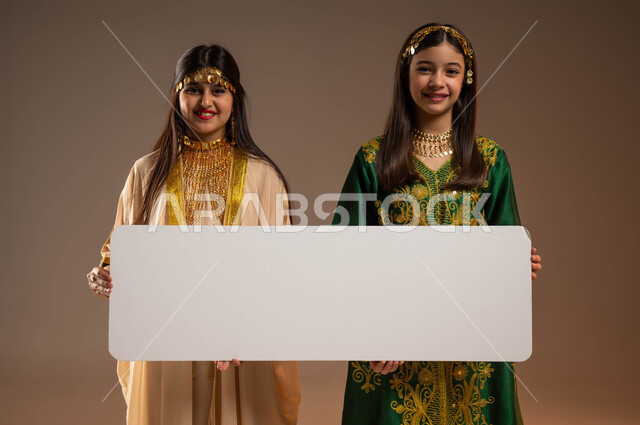 النظر إلى الكاميرا بإيماءات السرور، يوم بدينا 1727 م، ارتداء أزياء المنطقة الشرقية والوسطى في الأعياد والمناسبات الوطنية، ذكرى تأسيس الدولة السعودية الأولى 22 فبراير، بورتريه لفتاتين عربيتين خليجيتين سعوديتين مبتسمتين تحملان موك أب أبيض فارغ، خلفية بيج