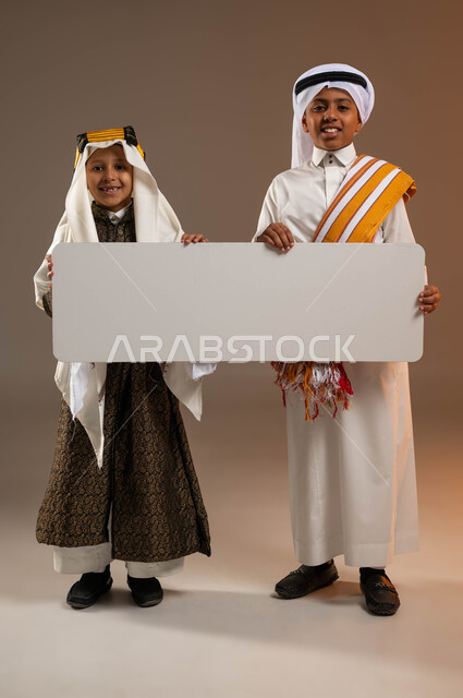 ذكرى تأسيس الدولة السعودية الأولى 22 فبراير، النظر إلى الكاميرا بإيماءات السرور، يوم بدينا 1727 م، ارتداء أزياء المنطقة الجنوبية والوسطى، بورتريه لولدين عربيين خليجيين سعوديين مبتسمين يحملان موك أب أبيض فارغ كبير، صورة لكامل الجسم، خلفية بيج