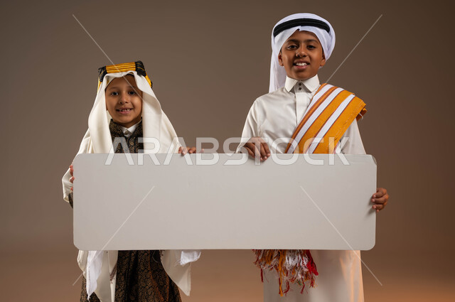 إحياء التراث السعودي، ذكرى تأسيس الدولة السعودية الأولى 22 فبراير، النظر إلى الكاميرا بإيماءات السرور، يوم بدينا 1727 م، ارتداء أزياء المنطقة الجنوبية والوسطى، بورتريه لولدين عربيين خليجيين سعوديين مبتسمين يحملان موك أب أبيض فارغ كبير، خلفية بيج