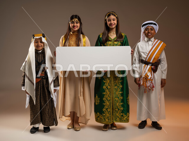 يوم بدينا 1727 م، ذكرى تأسيس الدولة السعودية الأولى 22 فبراير، النظر إلى الكاميرا بإيماءات السرور، ارتداء أزياء المنطقة الجنوبية والوسطى، بورتريه لمجموعة أولاد وبنات عرب خليجيين سعوديين مبتسمين يحملون موك أب أبيض فارغ كبير، صورة لكامل الجسم، خلفية بيج