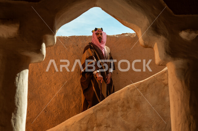 ارتداء الأزياء الشعبية في الأعياد والمناسبات الوطنية, الوقوف بفخر وشموخ في أحد البيوت والأحياء التراثية الطينية القديمة في النهار, ذكرى تأسيس الدولة السعودية الأولى 22 فبراير, رجل عربي خليجي سعودي يرتدي البشت والعقال الذهبي يرتكز على عصا خشبية, يوم بدينا 1727م