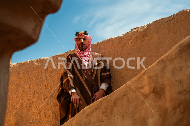 الوقوف بفخر وشموخ في أحد البيوت والأحياء التراثية الطينية القديمة في النهار, ذكرى تأسيس الدولة السعودية الأولى 22 فبراير, رجل عربي خليجي سعودي يرتدي البشت والعقال الذهبي يرتكز على عصا خشبية, يوم بدينا 1727م، ارتداء الأزياء الشعبية في الأعياد والمناسبات الوطنية