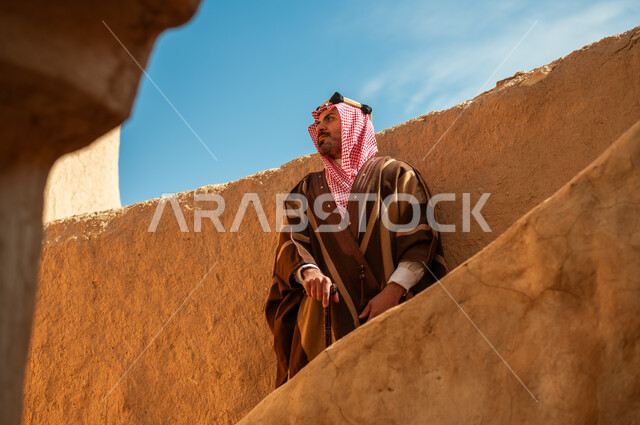 ذكرى تأسيس الدولة السعودية الأولى 22 فبراير, رجل عربي خليجي سعودي يرتدي البشت والعقال الذهبي يرتكز على عصا خشبية, يوم بدينا 1727م، ارتداء الأزياء الشعبية في الأعياد والمناسبات الوطنية، الوقوف بفخر وشموخ في أحد البيوت والأحياء التراثية الطينية القديمة في النهار