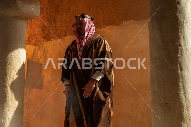 ارتداء الأزياء الشعبية في الأعياد والمناسبات الوطنية، الفخر والاعتزاز بتراث الوطن، ذكرى تأسيس الدولة السعودية الأولى 22 فبراير, رجل عربي خليجي سعودي يرتدي البشت والعقال الذهبي يقف في أحد البيوت والأحياء الأثرية الطينية القديمة في السعودية, يوم بدينا 1727 م