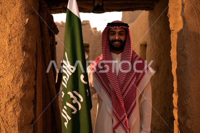 النظر الى الكاميرا بتعابير الاعتزاز والفخر بإنجازات الوطن، شاب عربي خليجي سعودي مبتسم يرتدي الثوب التقليدي والشماغ يمسك بيده علم التأسيس، الوقوف في إحدى الأحياء والبيوت الطينية التراثية القديمة, يوم بدينا 1727 م, ذكرى تأسيس الدولة السعودية الأولى في 22 فبراير