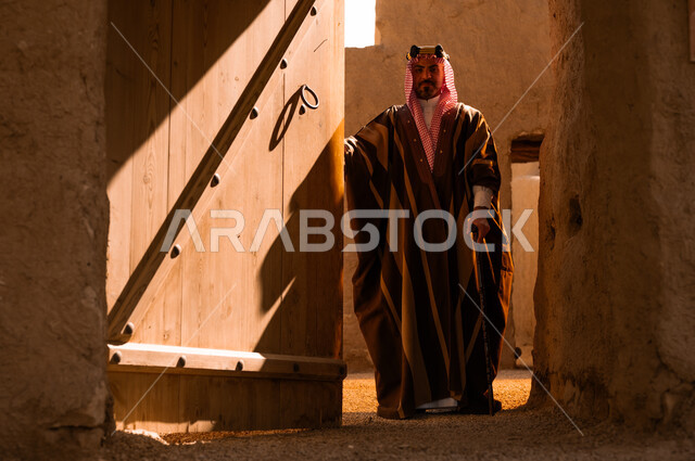 ارتداء الأزياء الشعبية في المناسبات الوطنية، ذكرى تأسيس الدولة السعودية الأولى في 22 فبراير، الأحياء والبيوت الطينية الاثرية التاريخية، رجل عربي خليجي سعودي يرتدي البشت والعقال الذهبي يقوم بفتح باب خشبي كبير، يوم بدينا 1727 م، الاعتزاز والفخر بأصالة الوطن