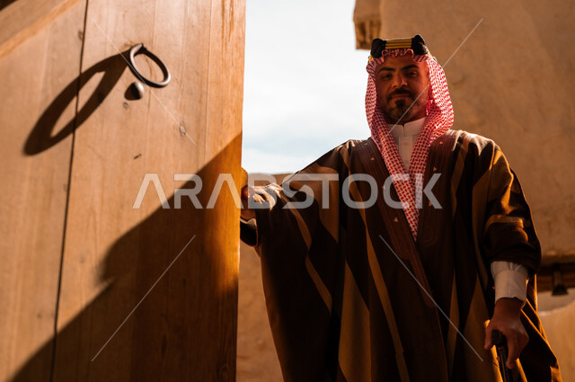 الاعتزاز والفخر بأصالة الوطن، ارتداء الأزياء الشعبية في المناسبات الوطنية، ذكرى تأسيس الدولة السعودية الأولى في 22 فبراير، الأحياء والبيوت الطينية الاثرية التاريخية، رجل عربي خليجي سعودي يرتدي البشت والعقال الذهبي يقوم بفتح باب خشبي كبير، يوم بدينا 172 م