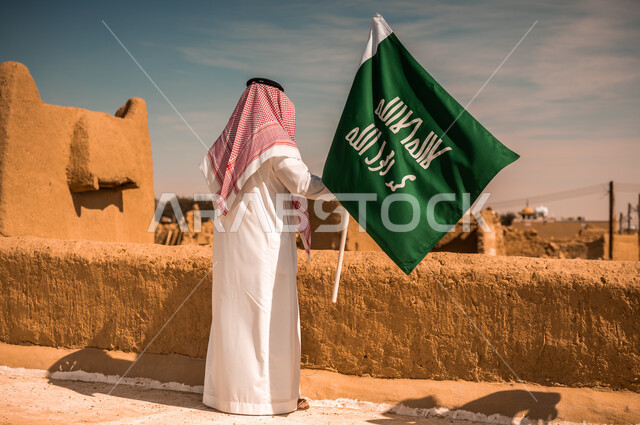 الوقوف باستقامة وبتعابير الاعتزاز والفخر بإنجازات الوطن، صورة من الخلف لشاب عربي خليجي سعودي يرتدي الثوب التقليدي والشماغ يمسك بيده علم التأسيس، الأحياء والبيوت الطينية التراثية القديمة, يوم بدينا 1727 م, ذكرى تأسيس الدولة السعودية الأولى في 22 فبراير