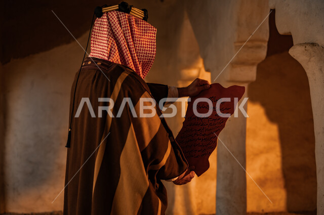 Preserving historical monuments and manuscripts, pride in the country’s authenticity and history, the anniversary of the founding of the first Saudi state, February 22, the Day of Dina 1727 AD, old mud heritage houses, a close-up photo from the back of a Saudi Gulf Arab man wearing a shemagh and bisht, reading letters from King Abdulaziz