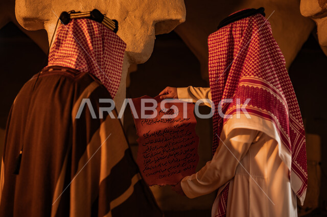 ذكرى تأسيس الدولة السعودية الأولى 22 فبراير، يوم بدينا 1727 م، توارث الأثار والمخطوطات بين أبناء الوطن، البيوت التراثية الطينية القديمة، صورة مقربة من الخلف لرجلين عربيين خليجيين سعوديين يرتديان الأزياء التقليدية الشعبية يقومان بقراءة رسائل الملك عبد العزيز