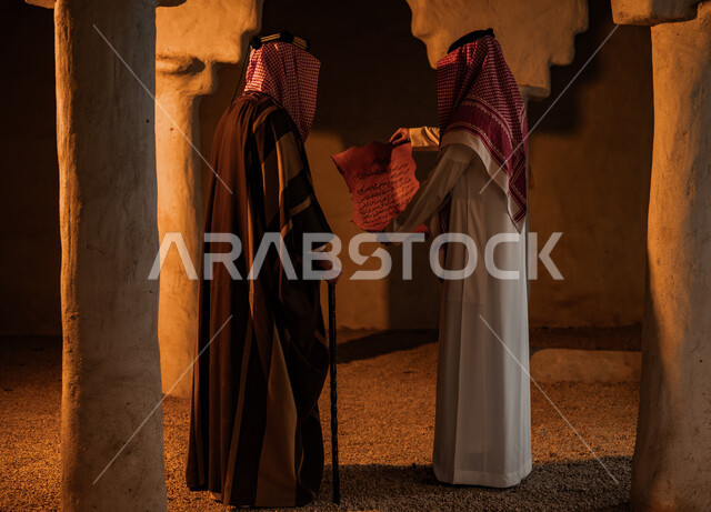 يوم بدينا 1727 م، ذكرى تأسيس الدولة السعودية الأولى 22 فبراير، توارث الأثار والمخطوطات بين أبناء الوطن، الأحياء والبيوت التراثية الطينية القديمة، صورة من الخلف لرجلين عربيين خليجيين سعوديين يرتديان الأزياء التقليدية الشعبية يقومان بقراءة رسائل الملك عبد العزيز