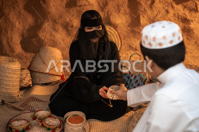 أعمال ومهن نسائية قديمة، وضع الحبوب في سلل من القش، منتجات ومحاصيل محلية وطنية سعودية، ذكرى تأسيس الدولة السعودية الأولى 22 فبراير، يوم بدينا 1727 م، صورة مقربة لإمرأة عربية خليجية سعودية ترتدي البرقع والعباءة تقوم ببيع البقوليات الى الولد في حي شعبي تراثي