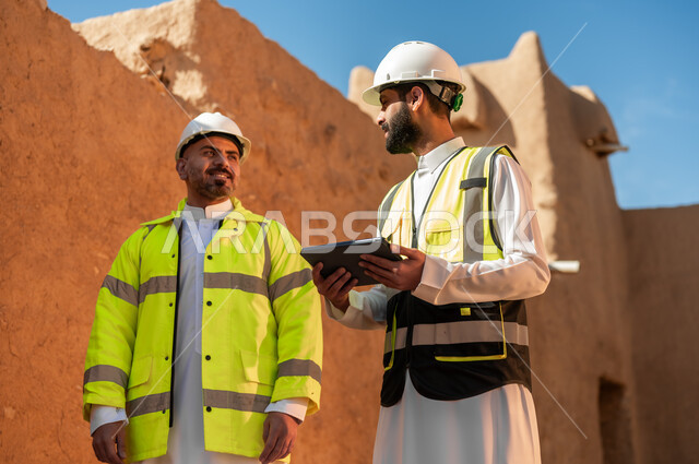 Using a tablet in engineering, the basics of building mud houses, discovering the ancient art of architecture, two Saudi Gulf Arab engineers wearing jackets and safety helmets standing on the ruins of Salwa Palace in Al-Bujairi, Diriyah, on our Day 1727 AD, the anniversary of the founding of the first Saudi state, February 22