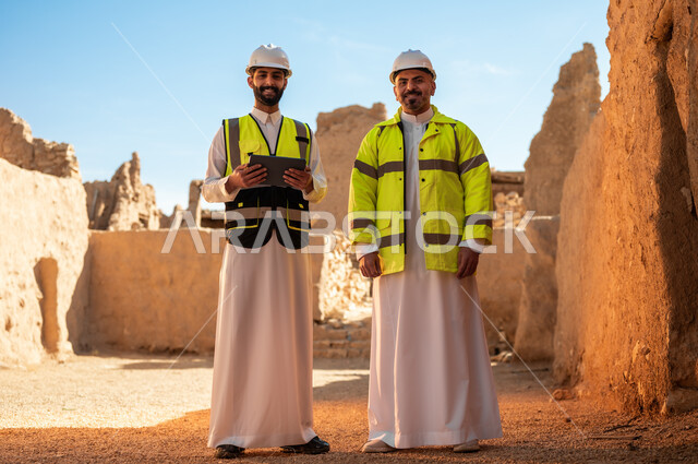 ذكرى تأسيس الدولة السعودية الأولى 22 فبراير، استخدام جهاز التابلت في الهندسة، اساسيات بناء البيوت الطينية، اكتشاف فن العمارة القديم، مهندسان عربيان خليجيان سعوديان يرتديان السترة وخوذة الحماية يقفان على اطلال قصر سلوى في البجيري الدرعية، يوم بدينا 1727 م