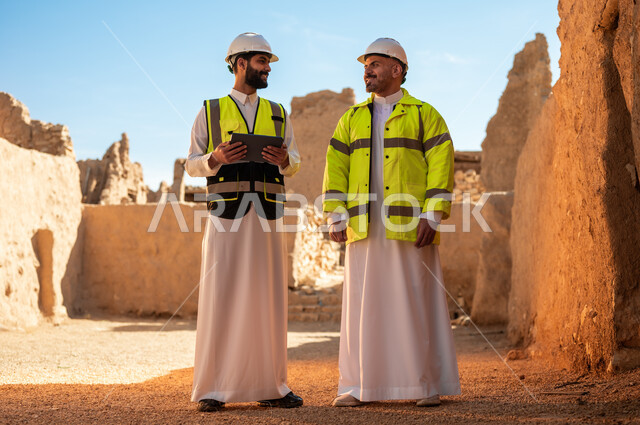 مهندسان عربيان خليجيان سعوديان يرتديان السترة وخوذة الحماية يقفان على اطلال قصر سلوى في البجيري الدرعية، يوم بدينا 1727 م، ذكرى تأسيس الدولة السعودية الأولى 22 فبراير، استخدام جهاز التابلت في الهندسة، اساسيات بناء البيوت الطينية، اكتشاف فن العمارة القديم