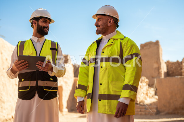 اساسيات بناء البيوت الطينية، اكتشاف فن العمارة القديم، استخدام جهاز التابلت في الهندسة،  مهندسان عربيان خليجيان سعوديان يرتديان السترة وخوذة الحماية يقفان على اطلال قصر سلوى في البجيري الدرعية، يوم بدينا 1727 م، ذكرى تأسيس الدولة السعودية الأولى 22 فبراير