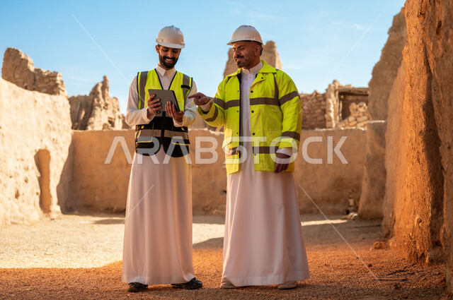 اساسيات بناء البيوت الطينية، اكتشاف فن العمارة القديم، مهندسان عربيان خليجيان سعوديان يرتديان السترة وخوذة الحماية يقفان على اطلال قصر سلوى في البجيري الدرعية، يوم بدينا 1727 م، ذكرى تأسيس الدولة السعودية الأولى 22 فبراير، استخدام جهاز التابلت في الهندسة