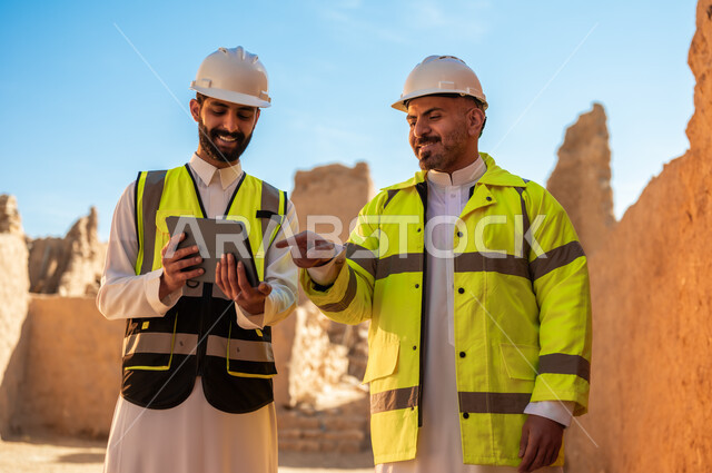 اكتشاف فن العمارة القديم، استخدام جهاز التابلت في الهندسة،  مهندسان عربيان خليجيان سعوديان يرتديان السترة وخوذة الحماية يقفان على اطلال قصر سلوى في البجيري الدرعية، اساسيات بناء البيوت الطينية، يوم بدينا 1727 م، ذكرى تأسيس الدولة السعودية الأولى 22 فبراير