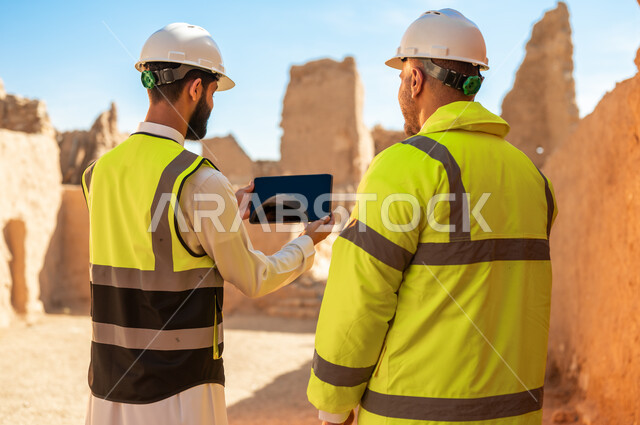 استخدام جهاز التابلت في الهندسة، فن العمارة القديم واساسيات بناء البيوت الطينية، صورة من الخلف لمهندسيين عربيين خليجيين سعوديين يرتديان السترة وخوذة الحماية على اطلال قصر سلوى في البجيري الدرعية، يوم بدينا 1727 م، ذكرى تأسيس الدولة السعودية الأولى 22 فبراير