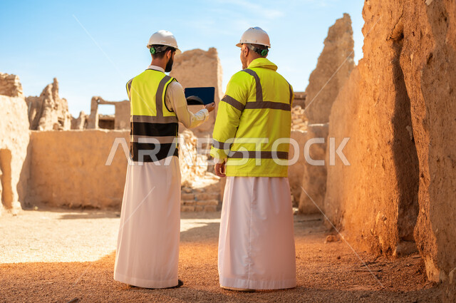  يوم بدينا 1727 م، ذكرى تأسيس الدولة السعودية الأولى 22 فبراير، استخدام جهاز التابلت في الهندسة، فن العمارة القديم واساسيات بناء البيوت الطينية، صورة من الخلف لمهندسيين عربيين خليجيين سعوديين يرتديان السترة وخوذة الحماية على اطلال قصر سلوى في البجيري الدرعية