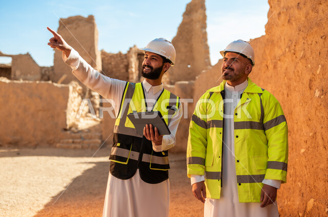 الاشارة الى شيء ما، اكتشاف فن العمارة القديم وبناء البيوت الطينية، مهندسان عربيان خليجيان سعوديان يرتديان السترة والخوذة يقفان على اطلال قصر سلوى في البجيري الدرعية، يوم بدينا 1727 م، ذكرى تأسيس الدولة السعودية الأولى 22 فبراير، استخدام جهاز التابلت في الهندسة