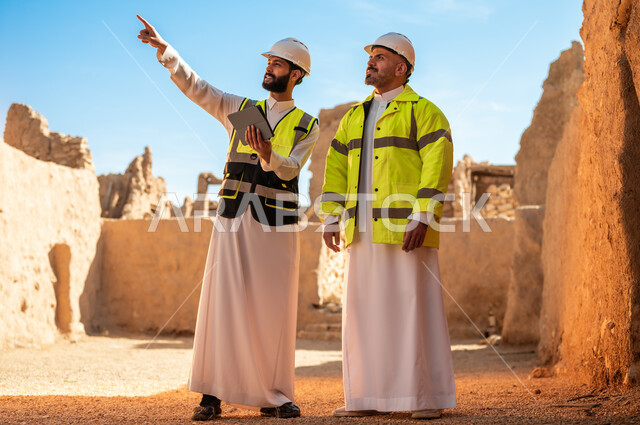 الاشارة الى شيء ما، اكتشاف فن العمارة القديم وبناء البيوت الطينية، مهندسان عربيان خليجيان سعوديان يرتديان السترة والخوذة يقفان على اطلال قصر سلوى في البجيري الدرعية، يوم بدينا 1727 م، ذكرى تأسيس الدولة السعودية الأولى 22 فبراير، استخدام جهاز التابلت في الهندسة
