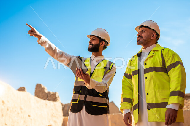  استخدام جهاز التابلت في الهندسة، الاشارة الى شيء ما، اكتشاف فن العمارة القديم وبناء البيوت الطينية، مهندسان عربيان خليجيان سعوديان يرتديان السترة والخوذة يقفان على اطلال قصر سلوى في البجيري الدرعية، يوم بدينا 1727 م، ذكرى تأسيس الدولة السعودية الأولى 22 فبراير